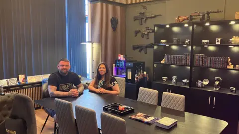 A man and woman are sat at one end of a long table, looking towards the camera. There are weapons mounted on the wall behind them, and a number of old cameras on the shelves