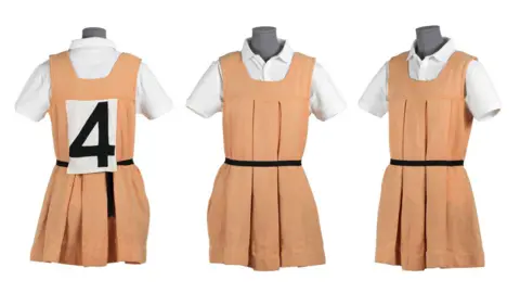 The National Museum of Ireland Three images of a camogie dress (front, back and side) on a mannequin. The dress is peachy orange in colour with a black belt and a white polo shirt underneath.