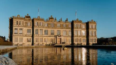 Tom Anders A large stately home with a square pond in front of it. It is a wintery setting with a blue sky behind the home.