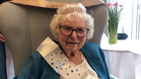 Betty Everest is wearing a crown and a sash to celebrate her 100th birthday.
Ms Everest is smiling at the camera while wearing a white blouse and a teal cardigan.