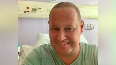 Jason Walker Jason sits in a hospital bed and wears a light green t-shirt. He has blue eyes and short shaved hair and is smiling.