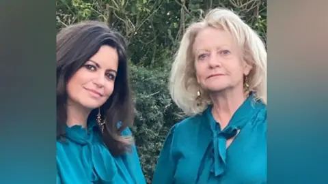 A picture of Dame Deborah James and her mum Heather James. Dame Deborah has brown hair and is wearing a blue shirt. Heather has blonde hair and is wearing a blue dress.
