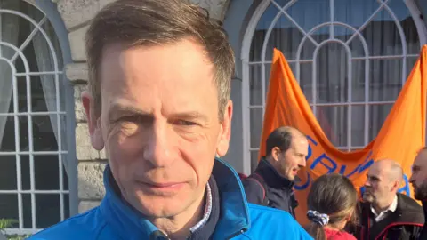 Jeremy Smith, with short greyish brown hair and wearing a blue coat, standing outside of a council building. A group of men and women are chatting behind him. 
