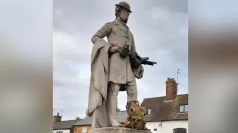 The statue of Lord John Scott, in Dunchurch, Warwickshire, stands on a plinth with houses in the background. It has been given military gear including an old British tin helmet, with a gun, flask, belt and rucksack.