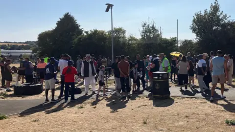 Louise Parry/BBC A crowd of children and adults mill about at the park, which is brown from the drought. There are a mix of different cultural backgrounds and some teenagers are using outdoor gym equipment.