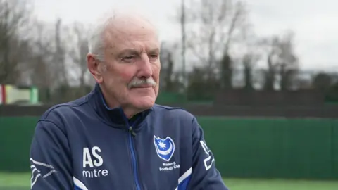 Alan Scott has short white hair and a white moustache. He is wearing a blue hoodie and is standing on a caged artificial football pitch.