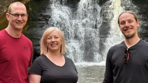 Two men and one woman - they look youngish in their 30s- standing in front of a waterfall.