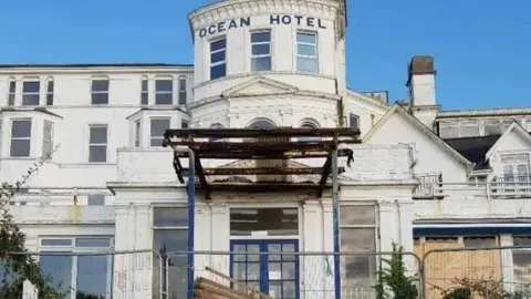 White building with multiple windows and curved entrance over three floors fencing and boards over some windows on the ground floor