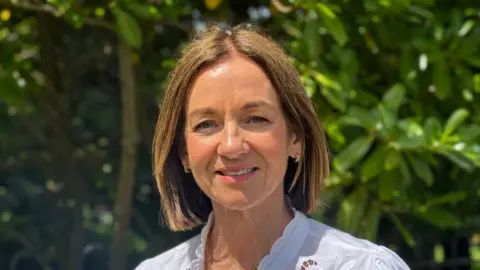 Councillor Gillian McCollum has short brown hair and is smiling at camera. Greenery in background.
