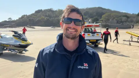 Tim wearing a navy RNLI Lifeguards jacket standing on St Brelade's beach. Behind him are rescue watercraft on trailers and an RNLI vehicle, with several other lifeguards walking nearby in wetsuits. A tree‑covered hillside rises in the background under a clear sky.
