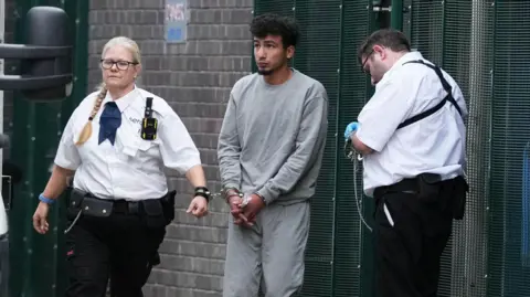 Eddie Mitchell A man in grey clothing and handcuffs is with two security officers beside a brick wall and metal fencing.