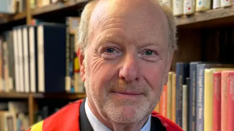 Sir Alan Bates sits in Bangor University library, wearing red, gold and black doctorate gown and a black suit, with a blue shirt and brown tie. Behind him are shelves of books.