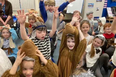 The picture shows excited children cheering and raising their hands as they get ready to perform their nativity show. Some are dressed as camels and Roman soldiers. 