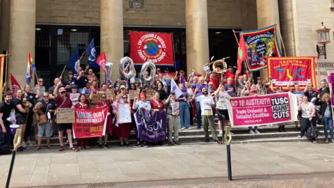 A large group of phlebotomists, union staff and supporters stand on the steps of Shire Hall in Gloucester on the picket line. They are holding banners, balloons, and signs calling for more pay and for their skills to be valued.