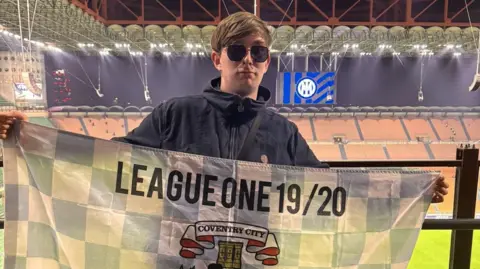 Alex Craig A man in sunglasses and a black coat is standing in a football stadium holding up a flag that reads League One 19/20 eith a Coventry City FC logo