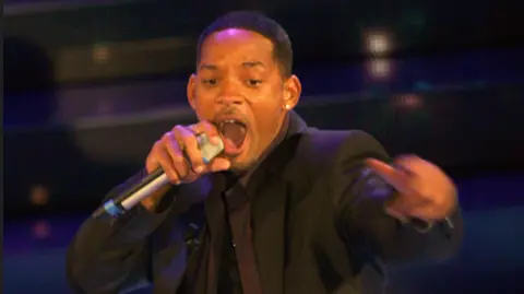 Getty Images Will Smith is holding a microphone and wearing a black suit on a stage.