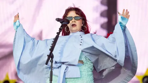 CMAT performs on Radio 1's Big Weekend stage against a pink backdrop with yellow stars. She wears a light blue floral two-piece outfit with a blue striped cape and has shoulder length red hair. She wears silver hoops and sunglasses and holds up her hands to the crowd