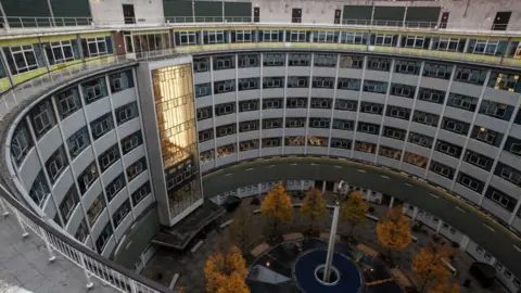 A U-shaped building with lots of dark windows and cladding, there are trees in the middle section and a large rectangular lift.