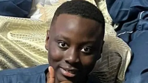 A smiling teenager sits and gives a thumbs up to the camera.