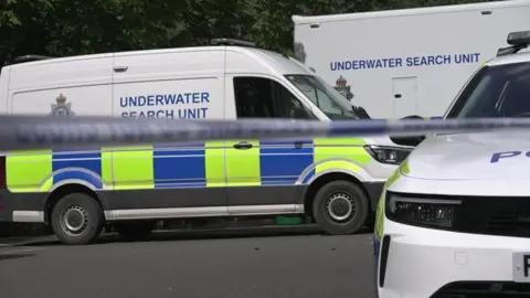 Three police vehicles. There which have Underwater Search Unit written on them. The vehicles are white with blue and yellow markings. 