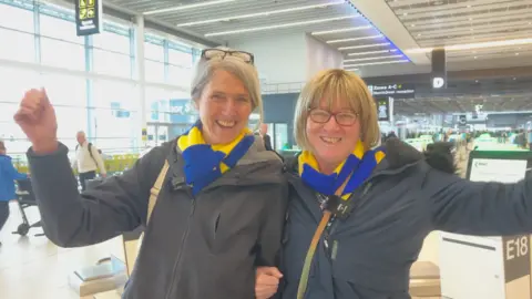 Carolyn (left) has her arm linked with Jeanette's. They are both wearing dark jackets and blue and yellow striped scarves. Carolyn has grey hair pulled back from her face and black glasses on her head. Jeanette has blonde bobbed hair, a fringe and wears dark-rimmed glasses.