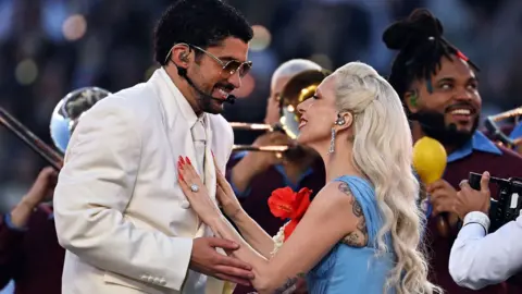 Bad Bunny with Lady Gaga on stage at the Superbowl. He is wearing a white suit and she is wearing a Blue dress . They are looking at each other and smiling