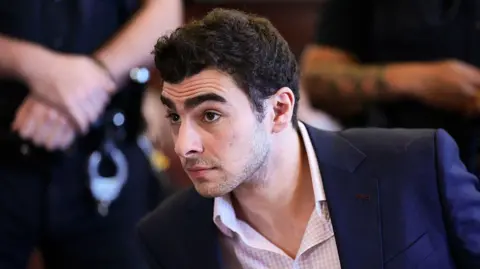 Luigi Mangione appears for a suppression of evidence hearing on 9 December 2025. He wears a pink checkered shirt and navy blue blazer.