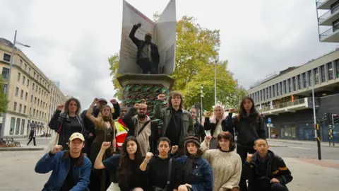 Plaster Creative Communications About a dozen young people standing in front of a statue on Colston plinth in Bristol - all of them raising a fist in the air and looking defiant. A large greetings card is balanced on the plinth.