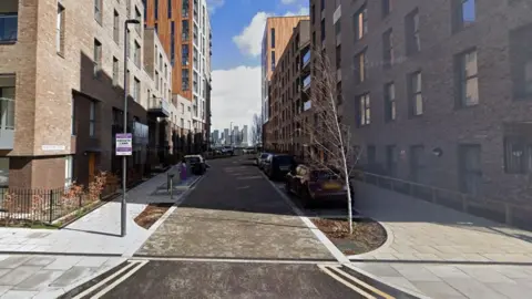 Google Maps New Union Close which shows high rise apartments on both sides, a paved road and cars parked in bays. In the far distance there is the London skyline of tower blocks and blue sky.