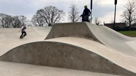 Three people on skateboards and helmets skating on a curved surface in a park.
