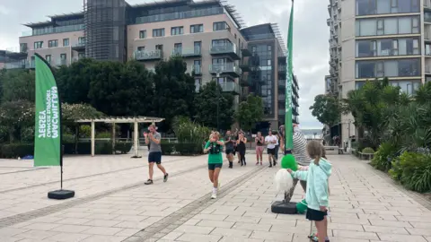 BBC Runners participating in an event on a paved urban pathway, flanked by modern buildings and greenery. Two green 'Macmillan Cancer Support' flags are visible on the left. A child in a light blue jacket stands on the right, cheering with white pom-poms. The sky is overcast