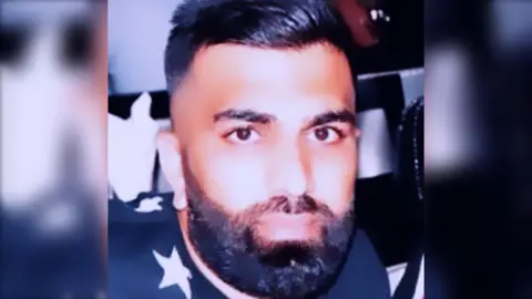 A man with short, dark hair and a short, dark beard looks at the camera