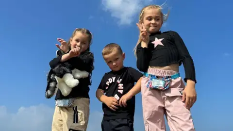 Siblings Paisley, Louis and Evie are standing side-by-side on a summer's day. The camera is pointing upwards, meaning that we don't see the ground, only the blue skies behind them. Paisley has her arms crossed in the air and is holding a cuddly toy. Evie is making a peace sign. Louis in the middle is staring directly at the camera. 