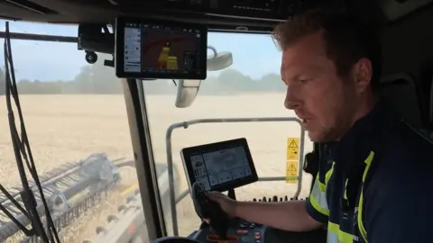 The camera operator is in the cab of a combine harvester being operated by Tom Hull. He is looking down at the controls as he harvests the wheat field.