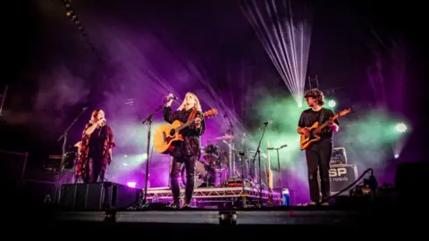 Tiree Music Festival Musicians perform on a festival stage with guitars and microphones, surrounded by purple, green and white stage lighting and light haze.
