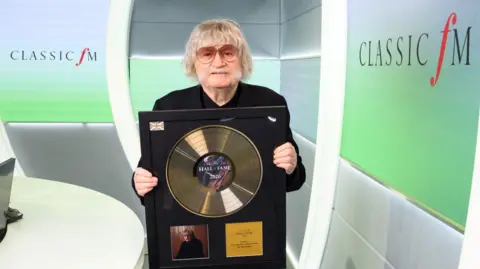 A grey haired man with a big grey moustache - wearing orange shaded sunglasses - holds up his Hall of Fame gold vinyl trophy. He stands in front of a branded screened booth, reading 'Classic FM'.