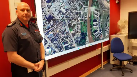 LDRS A man with a shaved head stands by a large TV screen on a wall showing a satellite mapping image of Shrewsbury town centre. He is in front of a red wall in an office environment.