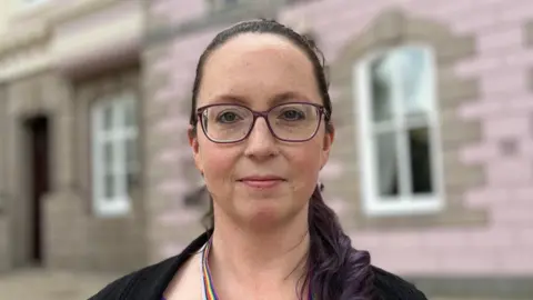 A woman called Louise Doublet stands in front of a pink building, which is blurred behind her. She has a neutral look on her face and is wearing large purple-rimmed glasses. She has long black hair tied into a ponytail and has a rainbow lanyard around her neck.