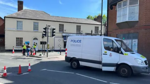 A police van and two police officers at the scene of the attack in June 2024. There is police tape across a town centre road.