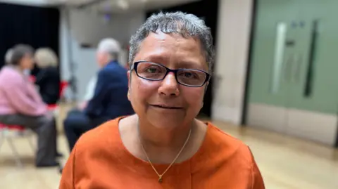A South-Asian woman with grey hair and glasses, wearing an orange shirt.