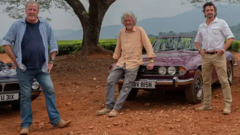 PA Jeremy Clarkson, James May and Richard Hammond filming the final The Grand Tour series on Amazon. They are stood together by cars on a dusty/clay surface outdoors with a large tree behind them. They're all wearing causal clothing.