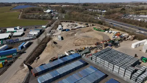 BBC A photo taken from a drone, showing a stretch of industrial land with cars and lorries parked on it. There's a road on the left hand side and a motorway on the right.