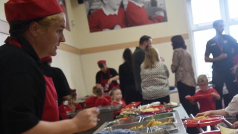 A woman dressed in a red and black catering uniform serving chips to schoolchildren