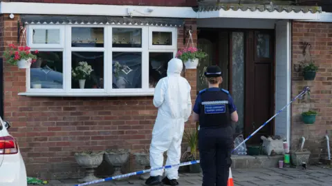 EDDIE MITCHELL A police community support officer and a forensic examiner in a white suit are outside a modern red brick property, which has been sealed off with blue and white police incident tape. 