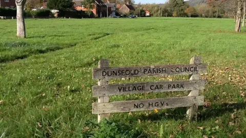 A sign on a green space near some houses in a village. The sign says 'DUNSFOLD PARISH COUNCIL, VILLAGE CAR PARK, NO HGVS.'