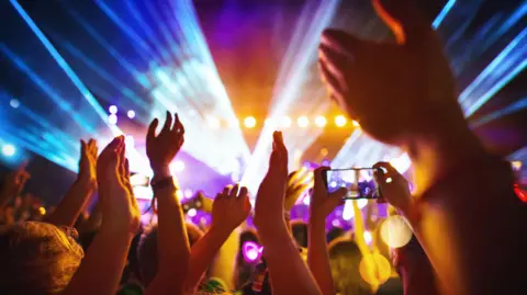 Rear view of a crowd of fans watching a DJ performance at a music festival. Rays of coloured lights are emitted from the stage. There are many raised hands, some are recording the show on mobile phones. 