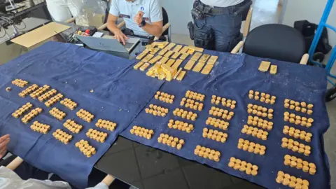 Lokale Politie Dendermonde Hundreds of gold coins and tens of gold bars laid out in stacks on a table covered with dark blue fabric. Two police officers can be seen sat at the table, with one on his laptop.