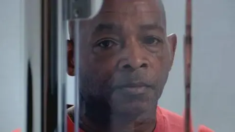 Channel 4 Man stares towards camera. He has a blank look on his face.