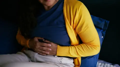 Getty Images A woman in a yellow jumper, blue t-shirt and grey jeans clutches her stomach. She is sitting on a bed in a dark room. Most of her face is not visible.