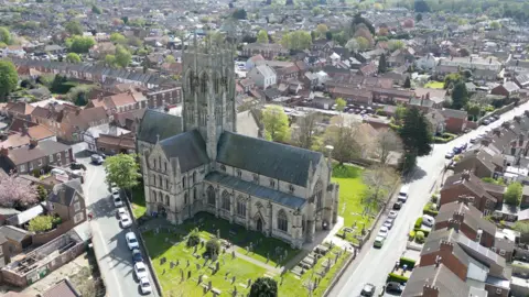 An aerial view shows a large historic stone church with a tall central tower, set within a graveyard and surrounded by residential streets. Rows of houses and tree‑lined roads extend around St Augustine's church.